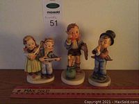 Photo showing 3 Hummel figurines in a row on wood shelf: boy with horn on green base, boy and girl with mandolins, and boy with art easel. Ruler shown for scale.