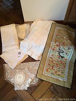 Overview of assorted linens including folded white and cream pieces with floral embroidery, multiple crocheted doilies, and an Asian-style silk runner.