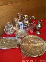 Overview of the lot on red tablecloth showing silver plated pitchers, candle holders, serving trays and plates wrapped in plastic
