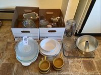 Baking dishes and kitchen utensils in two boxes and arranged on floor including glass, ceramic dishes, cups, and utensils.