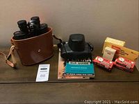 Photo showing Marksmann binoculars in brown leather case, Kodak films, and several camera manuals and guides on a wooden surface.