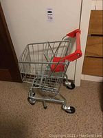 Full side view of the kid's metal shopping cart showing red handle and black wheels on a patterned floor.