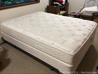 Full view of a white queen size mattress and box spring on a metal frame with wheels in a bedroom.