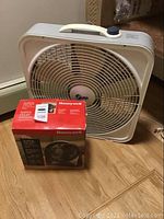 Large white Supertech box fan with handle and grill, shown on floor near baseboard heater