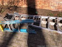Three aluminum ladders stacked outdoors on paver surface