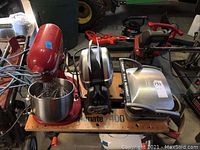 Three appliances displayed on wooden plank: red KitchenAid stand mixer with stainless steel bowl, silver waffle maker with stand behind Cuisinart griddler.