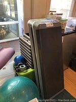 Photo showing two gray aerobic step platforms stacked against a window, with other exercise equipment nearby.