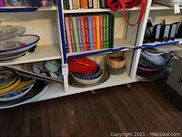 Wide shot of bottom shelf showing assorted serving bowls, platters, discs and wooden bowl and lidded pot with plastic bowls stacked to the right.