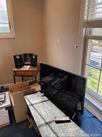 Photo showing 32 inch Vizio TV on a table near window with two remotes on table and a Philips Micro Hi-Fi system on small wooden side table with two satellite speakers and manuals.