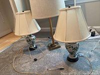 Photo of two cloisonné style lamps with white fabric shades and a taller faux brass finish floor lamp with a white fabric shade on carpet near wall.