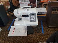 Wide shot showing Singer Curvy sewing machine model 8770 on carpet with instruction manual and foot pedal beside it