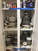 Photo of cabinet shelves containing stacked Studio Nova Impulse dishes, showing plates, bowls, cups, and other pieces with colorful geometric design.
