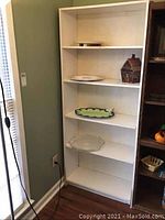 Front view of white particle board shelving unit showing multiple shelves with various items not included and visible scratches.