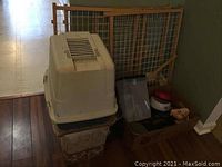 Photo showing a beige covered litter box on top of a patterned woven basket with two wooden pet gates behind, as well as pet dishes and other supplies on the floor.