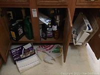 Cleaning supplies including Swiffer Wet Jet pads, Easy Off, Fabuloso, Cascade soap, furniture care kit inside a wooden cabinet along with plastic bags and bottles.