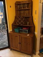 Photo showing full view of wooden corner cabinet placed in room corner. Upper shelving section with glassware and wine rack visible. Lower storage cabinet with double doors closed.