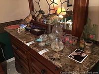 Wide view showing Royal Copenhagen crystal candle holder, oil lamp, reptile figurines including a turtle bank, metal tree jewelry box, small glass container with floral design, and assorted collectibles placed on a marble dresser top with wooden mirror.