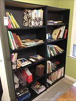 Books and magazines on multiple shelves of black bookcases, some books are standing upright while others are stacked or in bags.