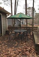 Full view of the outdoor patio set showing round metal table with glass top, four metal chairs, and green umbrella on a backyard deck with leaf-covered floor.