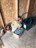Photo showing various flower pots, garden hose in container, bags and box with garden staples or stakes on floor against wall
