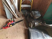 Photo of backyard tools including a push lawnmower with attached grass catcher bag and an electric chainsaw on a plywood floor.