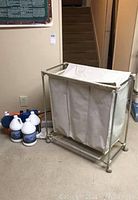 Laundry hamper with metal frame and three cream fabric compartments, multiple jugs of cleaning liquids nearby.
