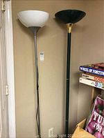 Two floor lamps standing side by side against a wall with neutral paint. One lamp features a white bowl-shaped shade, the other a black shade. Visible part of a shelf with board games is nearby.