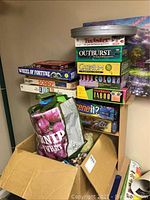 Stack of classic board games including Twister, Outburst, Cranium, True Colors, Celebrate Taboo, Scene It?, Wheel of Fortune, and Sorry stacked on a cabinet next to a cardboard box containing bags of toys.