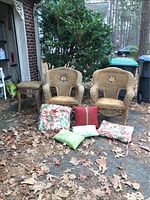 Three wicker furniture pieces arranged outside on driveway with dry leaves and some greenery visible. Two chairs and one side table with six cushions in front.