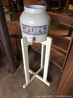 Full view of the ceramic drink crock on its white wooden stand showing the entire setup and surrounding area.