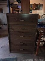 Front view of wooden chest of drawers showing five drawers with metal handles, dark brown wood finish, visible wear and scratches.