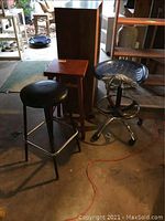 Photo showing three stools: black padded bar stool with chrome legs, wooden square stool, and metal tractor-style stool with contoured seat in garage setting.