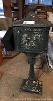 Full view of mailbox on pedestal with side mail retrieval door open showing ornate embossed design and chipped paint.