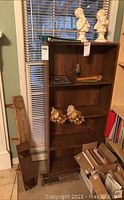 Wooden bookcase with three shelves, some decorative items on it, tags 546 and 547 visible on the furniture, window with blinds in the background.