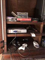 Wooden cabinet with PlayStation console and controllers on lower shelf, Sega Genesis and PlayStation games on upper shelves.