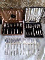 Two open boxes of silverplate fork sets and a row of forks laid out in front, showing three separate sets of forks with decorative handles and original box linings