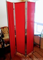 Photo showing the three-panel red fabric room divider standing fully unfolded in a room, metal frame visible along panel edges, fabric vibrant red and taut.