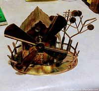 Front-left angle showing large four-blade propeller, antiqued tin finish, small roofed mill house, tree with round leaves, and fence on oval base.