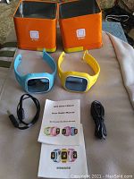 Photo showing two kids GPS smart watches in blue and yellow with black screens, two USB charging cables, user guide manual, and two orange metal storage boxes.