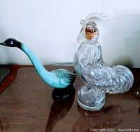Front angled view of a light blue and black pottery swan figurine beside a clear glass rooster decanter with amber-colored details on the head, placed on a wooden surface.