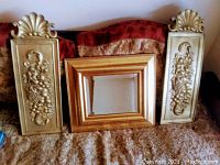 Overall photo of the two gold-coloured chalkware plaques with grape motif positioned on either side of a gold-toned framed mirror, all resting on a fabric surface.