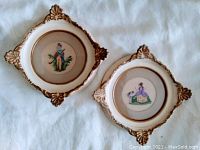 Two square cream and gilt framed needlepoint miniatures on white background. Both feature centered circular needlepoint designs with tan mattes.