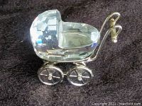 Side view of clear faceted Swarovski crystal baby carriage with silver metal frame and wheels, on dark fabric background.