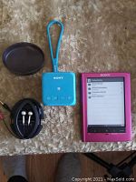 Photo showing pink Sony e-reader, blue Sony remote speaker with wrist strap, and black earbuds in black round case on textured surface.