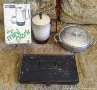 Complete lot including coffee percolator with box, aluminum serving dish with lid, and black metal antique cash box on a fabric surface.