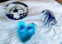 Three art glass items on white fabric: a blue and purple ashtray with white flowers, a blue glass Love Birds paperweight, and a clear and blue swirl glass paperweight.