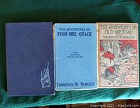 Photo showing the three vintage children's books laid side by side. From left to right: a dark blue book, a light blue book titled The Adventures of Poor Mrs. Quack, and a beige and red illustrated cover titled The Adventures of Old Mr. Toad by Thornton W. Burgess.