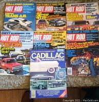 Lot of six vintage Hot Rod magazines from 1970s with colorful retro covers and one 1980 Cadillac Standard of Excellence hardcover book, all arranged on a surface for display.