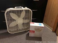 White box fan with metal grille and four blades, two control dials on top, shown on carpeted floor against dark background