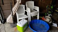 Set of outdoor items including 5 white plastic arm chairs stacked in the back, beige market umbrella with stand, green and white Coleman cooler, and blue plastic kiddie pool. All show outdoor use with dirt and weathering.